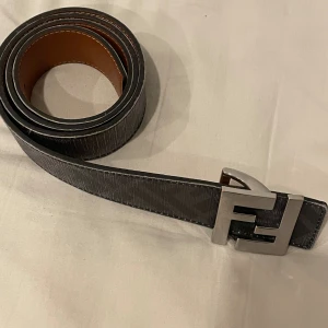 Svart Fendi bälte med FF-spänne - Säljer ett svart bälte från Fendi med klassiskt FF-mönster längs hela remmen och ett stort, blankt metallspänne i silver med Fendi-loggan. Bältet har en ganska stor repa i lädret, typ 5cm och färgen har repats bort till viss del