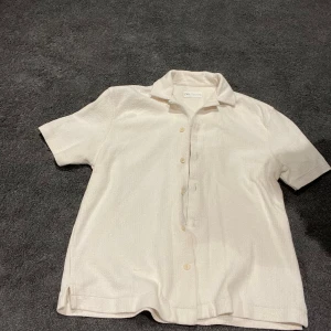 Vit kortärmad bomulls skjorta från Zara - Snygg vit bomulls skjorta från Zara med korta ärmar och klassisk krage. Skjortan har knappar framtill och är tillverkad i ett lätt bomullsmaterial som känns fräscht och luftigt. Perfekt för varma dagar och enkel att matcha med jeans eller shorts.