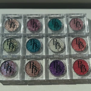 Glittriga ögonskuggor i 12 färger - Säljer ett set med 12 glittriga ögonskuggor i singelförpackningar. Färgerna är bland annat rosa, turkos, lila, guld, silver, grön och koppar. Skuggorna har intensivt skimmer och passar perfekt för kreativa sminkningar. Kommer i transparenta fyrkantiga dosor med logga på locket. 4A och A16 är SLUTSÅLDA! 60kr/st