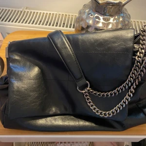 Svart axelväska med kedja från Zara - Snygg svart axelväska från Zara i skinnimitation med grova silverfärgade kedjor som axelrem. Väskan har ett stilrent, slätt yttre och praktiskt lock. Inuti finns dragkedja för extra förvaring. Perfekt för dig som gillar en cool och modern look.