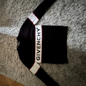 Svart stickad tröja från Givenchy - Svart stickad tröja från Givenchy med vita och röda detaljer. Tröjan har breda vita ränder med röda kanter på ärmarna och ett stort Givenchy-tryck över bröstet. Långärmad och med rund hals, perfekt för en snygg och trendig look.