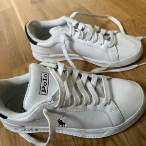 Vita sneakers från Polo Ralph Lauren - Snygga vita sneakers från Polo Ralph Lauren med klassisk design. Skorna har vit ovandel i skinn, vit sula och snörning. På sidan finns Polo-loggan broderad i svart. Insidan är fodrad i textil och yttersulan är i gummi. Perfekta för en clean och stilren look.