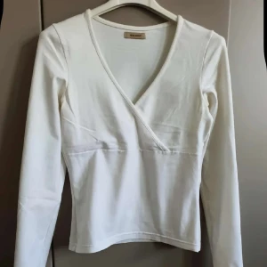 Vit omlottopp från Vero Moda - Snygg vit omlottopp från Vero Moda med lång ärm och v-ringning. Toppen har en figurnära passform och är tillverkad i mjuk bomull, perfekt att styla med jeans eller kjol. Enkel och stilren design som funkar året runt.