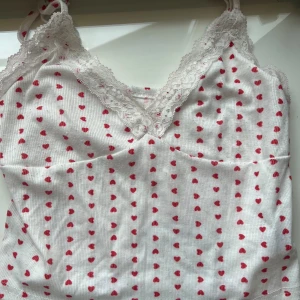 Vit hjärtmönstrad spetslinne från SHEIN - Supersöt vit linne från SHEIN med små röda hjärtan över hela tyget. Toppen har smala axelband och v-ringning med vit spetskant som ger en gullig vibe.             Längd=27cm 