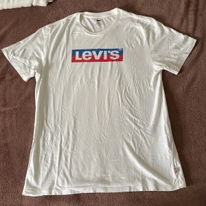 Vit Levi's t-shirt med logga, storlek L - Vit t-shirt från Levi's i storlek L med klassisk röd och blå logga på bröstet. T-shirten har rund hals och korta ärmar, tillverkad i mjuk bomull. Perfekt för en clean och avslappnad stil.