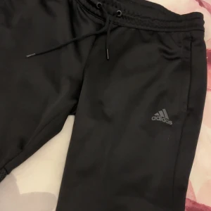 Svarta Adidas träningsbyxor med knappar - Svarta träningsbyxor från Adidas med klassiska vita ränder längs sidorna och tryckknappar hela vägen ner. Byxorna har snörning i midjan och meshdetaljer på sidorna för extra ventilation. Perfekta för sport eller chill dagar.