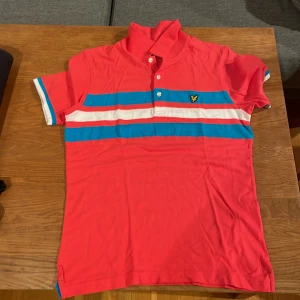 Randig pikétröja från Lyle & Scott - Säljer en snygg pikétröja från Lyle & Scott i medium. Tröjan är korallröd med breda blå och vita ränder över bröstet och ryggen. Klassisk krage, kortärmad och med knappar framtill. Ikonisk gul logga på bröstet. Perfekt för vår och sommar!