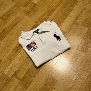 Vit pikétröja Ralph Lauren USA - Vit pikétröja från Ralph Lauren med broderad USA-flagga och text på bröstet samt stor polospelare-logga. På ryggen finns siffran 3, texten UNITED STATES och en till flagga. Tröjan är i custom fit och tillverkad i bomull med lite spandex för stretch.