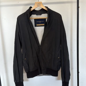 Svart bomberjacka från Henri Lloyd - Svart bomberjacka från Henri Lloyd med klassisk krage, ribbade muddar och dragkedja framtill. Jackan har två fickor med knappstängning och diskret broderad logga på bröstet. Perfekt för dig som gillar stilren och sportig look.