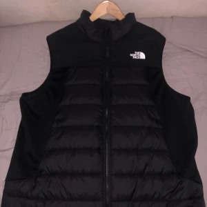 Svart dunväst från The North Face - Svart dunväst från The North Face med klassisk puffer-design och hög krage. Västen har en tydlig logga på bröstet och dragkedja framtill. Materialet är dun och polyester. Aldrig använd förut. Helt ny. Säljer för att den inte passade mig. Priset är inte hugget i sten.