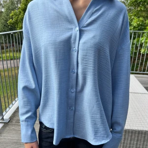 Ljusblå oversized skjorta med knappar från vero Moda - Snygg ljusblå skjorta med avslappnad oversized passform och klassisk krage. Skjortan har långa ärmar, knäppning framtill och är tillverkad i ett mjukt, lätt material som ger en skön känsla. Perfekt för en chill och stilren look.