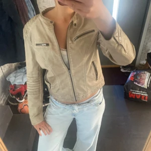 Beige skinnjacka med dragkedjor - Snygg beige skinnjacka med coola detaljer som dragkedjor på bröstet och vid fickorna. Jackan har rundad krage utan kragslag och markerade sömmar på axlar och ärmar. Den är lite sönder där bak men inget som gör nåt. Inte äkta skinn