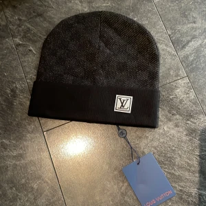 Svart mössa med logga - Snygg svart mössa med diskret rutigt mönster och vikt kant. Mössan är tillverkad i mjukt material och passar perfekt för kalla dagar när du vill ha en stilren look.