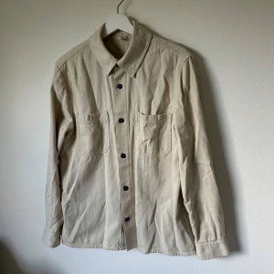 Beige overshirt från Arket i bomull - Stilren beige overshirt från Arket i ekologisk bomullstwill. Skjortan har två stora bröstfickor, svarta knappar och klassisk krage. Perfekt lager-på-lager-plagg med lång ärm och enkel, clean look. 2H15