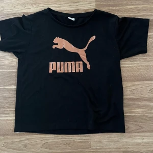 Svart Puma t-shirt med koppartryck - Svart t-shirt från Puma med stort kopparfärgat logotryck på bröstet. Klassisk rund hals och korta ärmar. Tillverkad i mjuk bomull som är skön mot huden. Perfekt för dig som gillar sportiga och stilrena plagg med tydlig branding.
