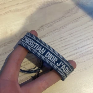 Blå Christian Dior J'Adior armband - Snyggt blått armband från Christian Dior med texten 'J'Adior' broderad i vitt. Armbandet har en bred, vävd design och avslutas med flätade snören och tofsar för justerbar passform. Perfekt statement piece för dig som gillar lyxiga accessoarer.