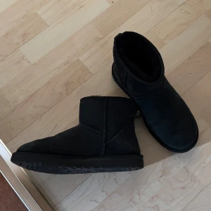 Svarta UGG boots i mocka - Mysiga svarta UGG boots i klassisk modell med rund tå och platt sula. Skorna är fodrade och tillverkade i mjuk mocka, perfekta för kalla dagar. UGG-logga bak på hälen och enkel design som passar till det mesta.