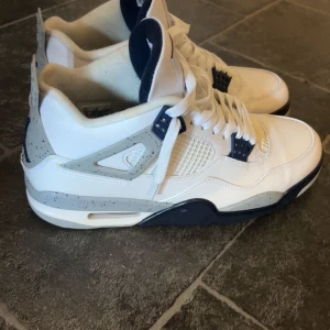 Nike Air Jordan 4  - Säljer ett par Nike Air Jordan 4 sneakers i vitt och marinblått med grå detaljer och stänkmönster. Skorna har klassisk snörning, meshpaneler och Jumpman-logga på plösen. Ovandelen är i skinn och sulan är vit med blå och grå inslag. Perfekta för dig som gillar streetwear.