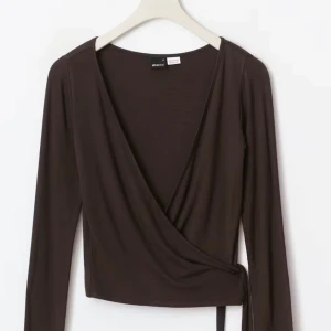 Brun omlottopp från Gina Tricot XS - Säljer en brun omlottopp från Gina Tricot i storlek XS. Toppen har lång ärm, djup v-ringning och knyt framtill för en snygg och trendig look. Tillverkad i mjukt bomullsmaterial som känns skönt mot huden. Perfekt att styla med jeans eller kjol.
