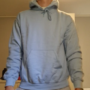 Ljusblå hoodie med tryck - Ljusblå hoodie med stor ficka framtill och huva med snörning. På ryggen finns ett stort tryck med en bodybuilder och texten 'MILHUNTER' i rött. Perfekt för dig som gillar unika och sportiga plagg.