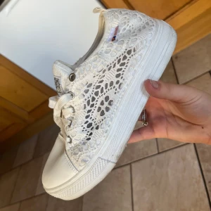 Vita spets-sneakers från OK Sport, stl 40 - Säljer ett par vita sneakers från OK Sport i storlek 40. Skorna har snygg spetsdetalj på sidorna, snörning framtill och platt sula. Perfekta för dig som gillar en feminin och fräsch stil. Märke och stjärndetaljer på plösen.