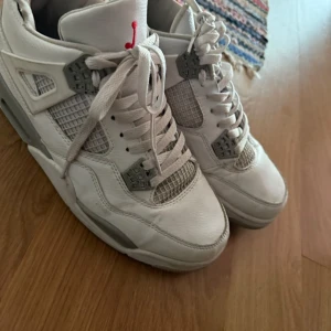 Nike Air Jordan 4 vita sneakers - Säljer ett par Nike Air Jordan 4 sneakers i vitt med gråa detaljer och röd logga på plösen. Skorna har klassisk snörning, meshpaneler och robust design. Perfekta för dig som gillar streetwear och vill sticka ut med en ikonisk modell.
