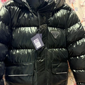 Svart pufferjacka från Prada, stl L - Säljer en svart, glansig pufferjacka från Prada i storlek L. Jackan har huva, stora fickor framtill och klassisk Prada-logga på bröstet. Tillverkad i syntetmaterial och perfekt för kalla dagar. Snygg och stilren design som sticker ut.