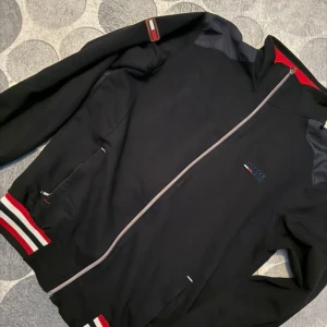 Svart bomberjacka från Tommy Hilfiger - Svart bomberjacka från Tommy Hilfiger med hel dragkedja, röda och vita detaljer på muddar och insida krage. Jackan har broderad logga på bröstet och klassisk sportig look. Perfekt för dig som gillar stilrena och snygga jackor.