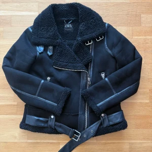 Svart teddyjacka från Zara med bälte - Säljer en svart teddyjacka från Zara med stor krage, dragkedja, bälte i midjan och detaljer i skinnimitation. Jackan har mysigt teddyfoder, långa ärmar och spännen vid kragen. Perfekt för kalla dagar.