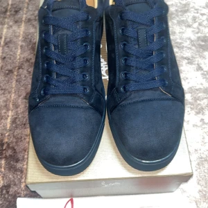 Christian Louboutin svarta sneakers mocka - Snygga svarta sneakers från Christian Louboutin i mocka med klassisk röd sula och broderad logga på hälen. Skorna har rund tå, platt sula och snörning. Perfekta för dig som vill ha en lyxig och stilren look med en touch av street.