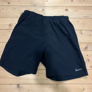 Svarta träningsshorts från Nike - Svarta träningsshorts från Nike med elastisk midja och diskret Nike-logga på benet. Tillverkade i lätt och snabbtorkande polyester, perfekta för sport eller gym. Vanlig passform med en innerbyxa som håller dom på plats. Pris kan alltid diskuteras vid snabb affär, postar samma dag.                                                              MVH