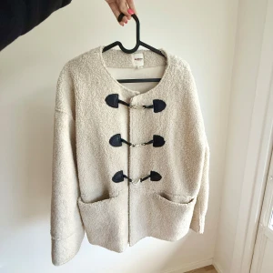 Beige teddyjacka med spännen från MQ - Supermjuk och oversized teddyjacka i beige från Dobber. Jackan har stora fickor framtill och coola svarta spännen som stängning. Perfekt för dig som vill ha en mysig och trendig look under höst och vinter.