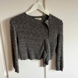 Grå ribbad kofta från Hollister - Mysig gråmelerad kofta från Hollister i ribbad stickad kvalitet. Modellen är croppad med långärmade ärmar och öppen framtill, perfekt att slänga över en topp. Snygg och enkel att matcha till olika outfits.