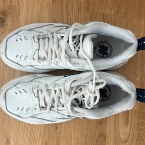 Vita New Balance sneakers med snörning - Säljer ett par chunky vita New Balance sneakers med blå detaljer och klassisk N-logga på sidan. Skorna har snörning, platt sula och rund tå. Ovandelen är tillverkad i skinn och syntet med ventilerande hål för extra komfort. Perfekt för dig som gillar retrostil och sportig vibe.