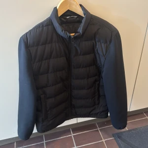 Svart pufferjacka med dragkedja - Svart pufferjacka från Zara Man med quiltad framsida och släta ärmar. Jackan har hög krage, hel dragkedja och två sidofickor. Perfekt för kyliga dagar och har en clean, stilren look som funkar till det mesta.