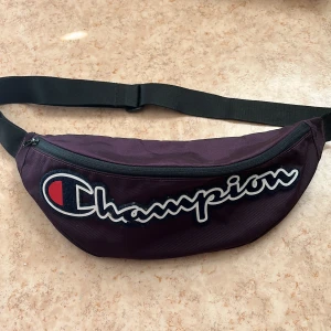 Mörklila magväska från Champion - Snygg mörklila magväska från Champion med stort broderat logotyptryck framtill. Väskan har justerbart svart band, dragkedja och innerfack med extra dragkedja.