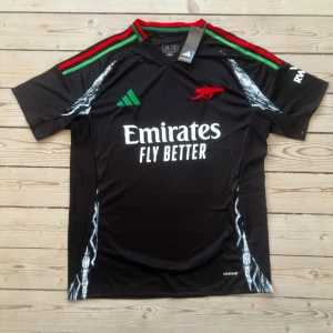 Arsenal svart fotbollströja Adidas 2XL - Svart Arsenal fotbollströja från Adidas i funktionsmaterial. Tröjan har gröna och röda ränder på axlarna, vit logga och tryck, samt detaljer i silver längs sidorna. Klassisk passform med korta ärmar och rund hals. Perfekt för dig som vill sticka ut på planen.