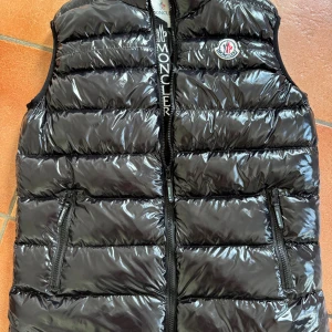 Svart dunväst från Moncler - Snygg svart dunväst från Moncler med glansig finish och klassisk logga på bröstet. Västen har två dragkedjefickor framtill, quiltad design och Moncler-tryck på dragkedjan. Perfekt för lager-på-lager och streetstyle.