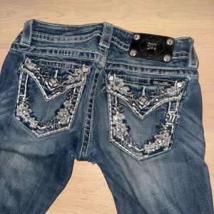Miss Me jeans med broderade bakfickor - Snygga blå jeans från Miss Me med broderade och dekorerade bakfickor med blommönster och strassdetaljer. Jeansen har kontrastsömmar, utsvängda/bootcut ben. Säljer pga att jag aldrig använder de då jag har ganska mycket jeans🩷 nypris när jag köpte var 1000kr och släjer för 590kr då framknsppen saknas, även är det lite hål vid skrevet samt lite slitningar nertill annars är de i bra skick. (Pris går att diskuteras) 25w. 