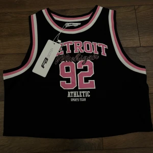 Svart sportig linnetopp - Basket inspirerad svart linnetopp från New Yorker med tryck 'Detroit Michigan 92' i rosa och vitt. Toppen har vita och rosa ränder längs halsringning och ärmhål, croppad passform och ärmlös design. Perfekt för en cool och avslappnad stil.