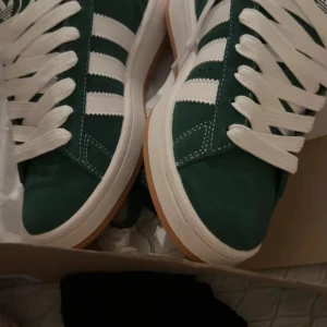 Adidas Campus gröna sneakers - Helt nya kommer med gröna snören oxå! Snygga Adidas Campus sneakers i mörkgrönt mocka med klassiska vita ränder och vit sula. Skorna har breda vita snören och en gummisula i beige. Perfekta för dig som gillar retrostil och streetwear.