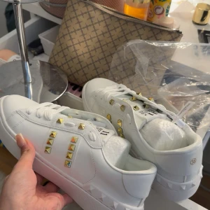 Valentino Rockstud vita sneakers - Snygga vita Valentino Rockstud sneakers i skinn med ikoniska guldiga nitar på sidorna och tungan. Klassisk rund tå, snörning och platt sula med mönstrad undersida. Perfekta för dig som vill sticka ut med en lyxig och edgy vibe.