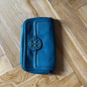 Blå axelväska från Tory Burch - Snygg blå axelväska från Tory Burch i skinn med guldfärgad kedjerem och ikoniskt logospänne framtill. Väskan har en stilren, rektangulär form och är perfekt för att bära det viktigaste. Kedjan kombinerar metall och skinn för en cool look.