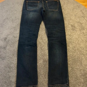 Mörkblå jeans från Nudie Jeans - Säljer ett par mörkblå jeans från Nudie Jeans med klassisk femficksdesign och kontrastsömmar. Jeansen har raka ben, subtila slitningar och Nudie-logga på bakfickan. Tillverkade i bomull och perfekta för en avslappnad stil.