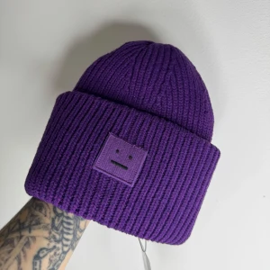 Lila mössa från Acne Studios - Snygg lila mössa från Acne Studios i grovstickad ull. Mössan har en bred uppvikt kant och ett ikoniskt ansiktsmärke framtill. Perfekt för dig som vill ha en cool och färgstark accessoar till vintern.