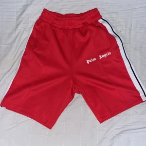 Palm Angels röda shorts med vit rand - Palm Angels shorts i röd färg med vit och svart rand längs sidorna. De har elastisk midja och två fickor. Loggan är tryckt i vitt på vänster sida. Perfekta för dig som gillar streetwear och sportig stil.