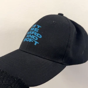 Svart Billie Eilish keps med blå text! - Svart keps med böjd skärm och trycket 'HIT ME HARD AND SOFT' i blått framtill. Klassisk modell med justerbar baksida, tillverkad i bomull. Perfekt accessoar för en cool och avslappnad stil.