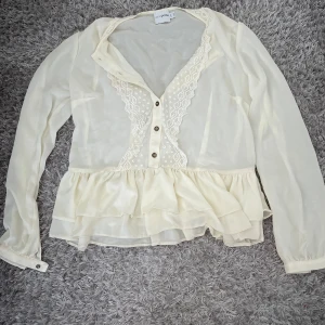 Vit blus med volang från ASOS Petite - Supersöt vit blus från ASOS Petite med volang nertill och spetsdetaljer längs v-ringningen. Blusen har knappar framtill och är gjord i ett tunt, lätt material som ger en luftig känsla. Långärmad och perfekt för dig som gillar romantisk stil.