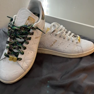 Adidas Stan Smith x BAPE sneakers - Unika Adidas Stan Smith x BAPE sneakers i vitt skinn med präglat mönster och gulddetaljer. Ena skon har vita snören, den andra har gröna camo-snören. Båda har guldiga 30 tals tags framtill för att representera bapes 30 års juvelerna. Kommer i original blå BAPE-box. Perfekt för dig som gillar limited edition och streetstyle och även grish  eller passar riktigt fet med ett par jeans. Bara ett par pytte små slitningar som säkert går bort om man tar ett våtservett o putsar lite 👍