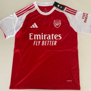 Arsenal Saka 7 matchtröja Adidas - Riktigt snygg Arsenal fotbollströja från Adidas med Saka och nummer 7 på ryggen. Tröjan är röd med vita ärmar och detaljer, klassisk klubbsköld på bröstet och Emirates Fly Better-tryck framtill. Tillverkad i lätt och ventilerande material, perfekt för match eller träning.